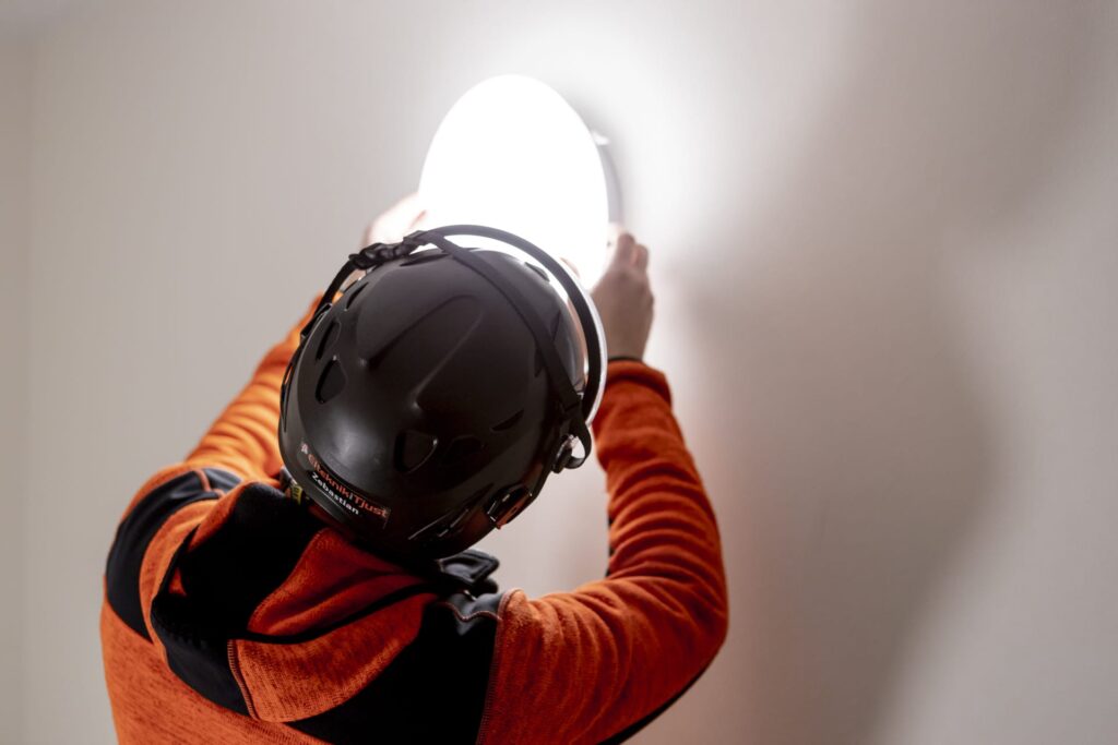 En elektriker med orange jacka och svart hjälm monterar en plafond lampa på en vägg.